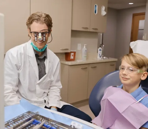 Dentist wearing magnification loupes smiling at a young patient seated in the dental chair during an exam