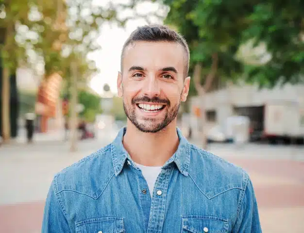 Smiling man with a beard in a denim shirt showing a bright, healthy smile outdoors