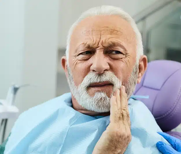 Older man in a dental chair holding his cheek and wincing from tooth pain