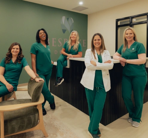 Dental practice team of five women in green scrubs and white coat posing together in modern office.