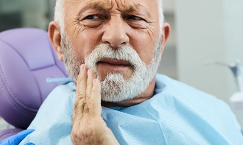Older man with beard experiencing tooth pain, holding hand to cheek in dental chair.