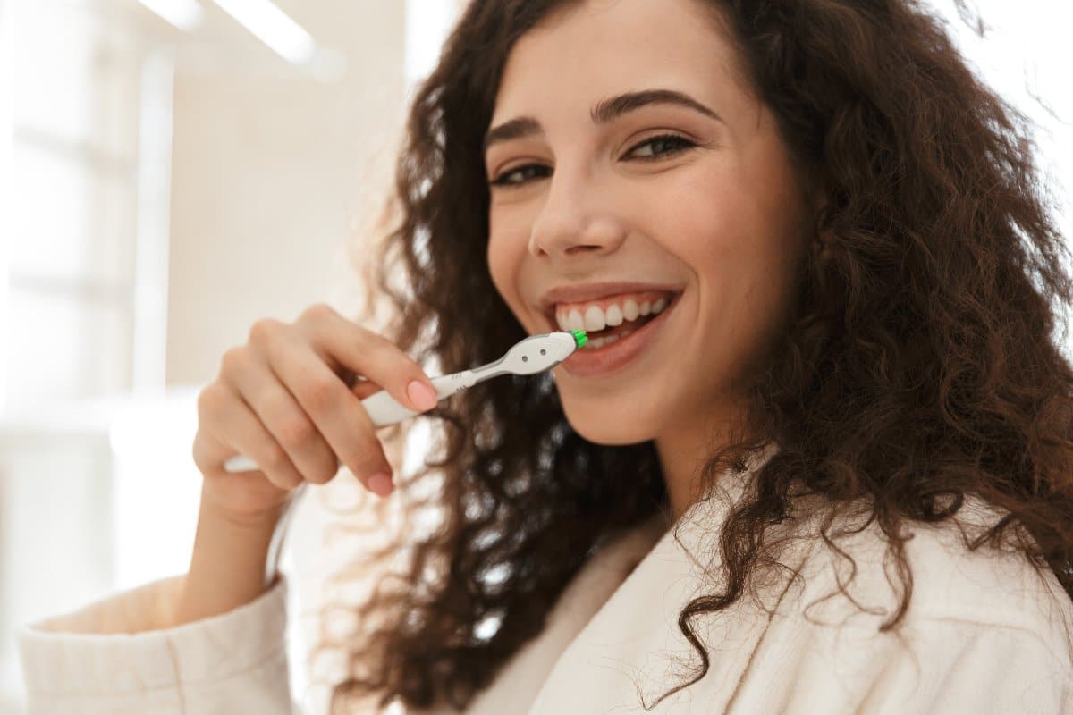 Woman brushing teeth at home, smiling at camera in bright bathroom setting.