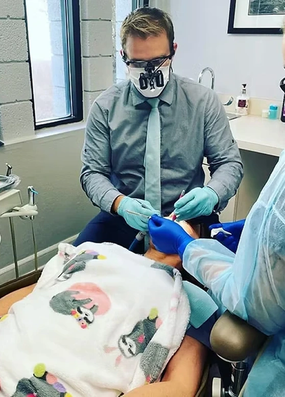 Dentist wearing surgical mask, gloves, and dental loupes actively performing a dental procedure on a patient in the office.