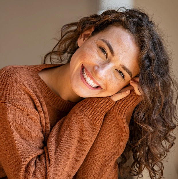 Young woman with curly hair and freckles smiling warmly while resting her head on her hand