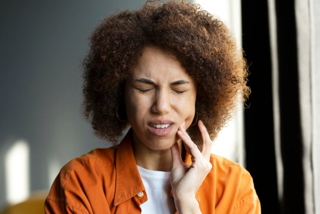 Young woman with natural hair holding her jaw in pain against an orange background
