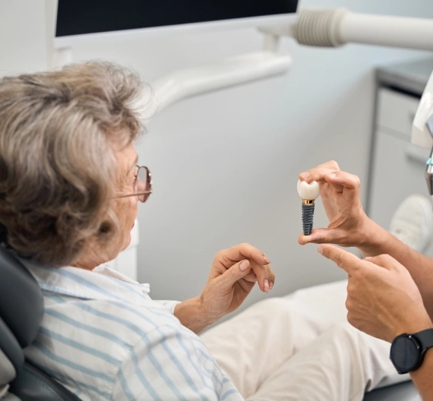 Un profesional de la odontología le muestra un modelo de implante dental a una paciente mayor sentada en el sillón dental.
