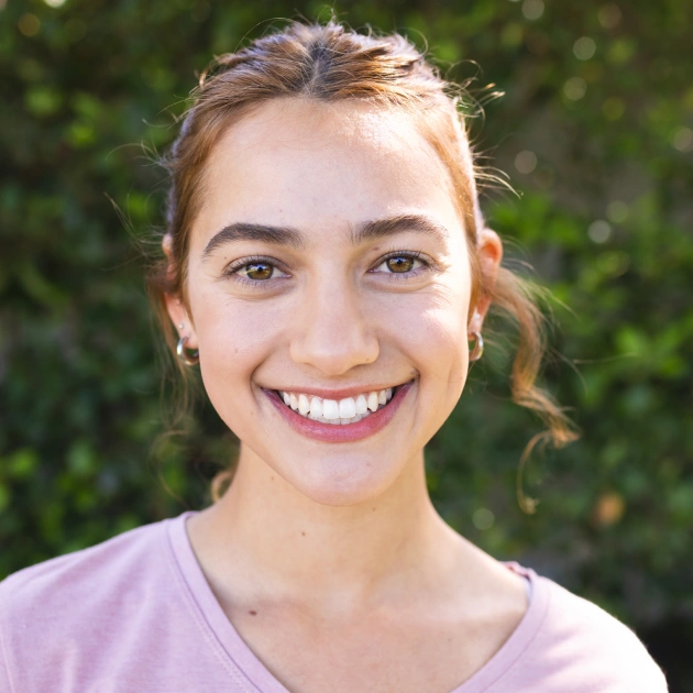 Retrato de una joven con cabello castaño rojizo recogido en un moño, sonriendo radiantemente al aire libre con vegetación de fondo.