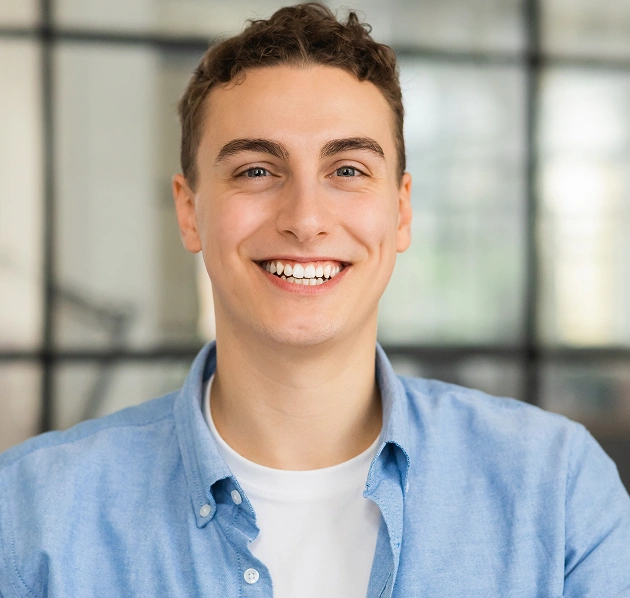 Portrait of a young man in a light blue shirt smiling broadly in a modern indoor setting