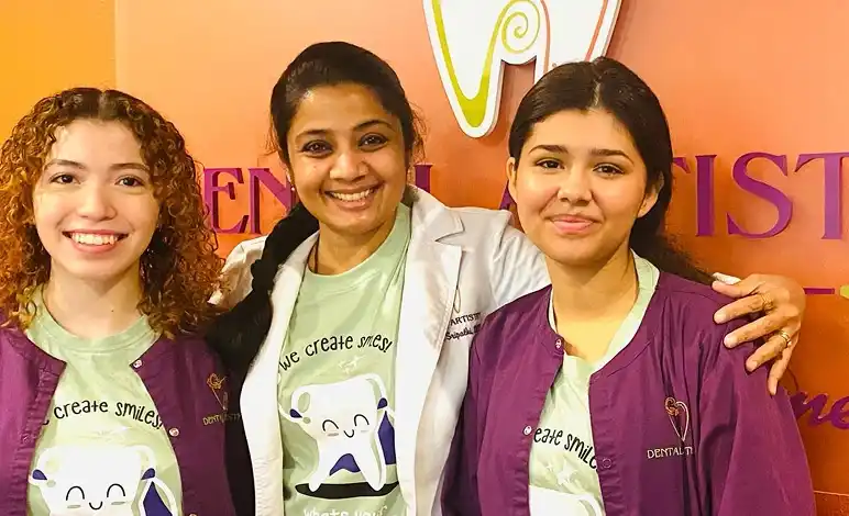 Dr. Radhika Srikrishnan Iyer and two Dental Artistry Irving team members wearing we create smiles shirts posing together in front of the office sign