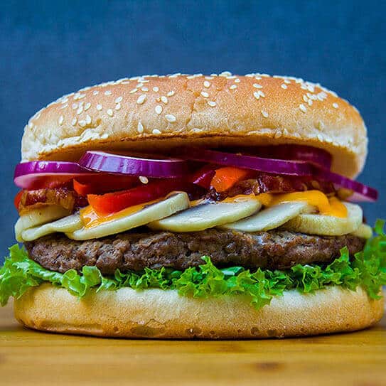 Gourmet burger with sesame seed bun, beef patty, lettuce, tomato, and purple onion on wooden surface.