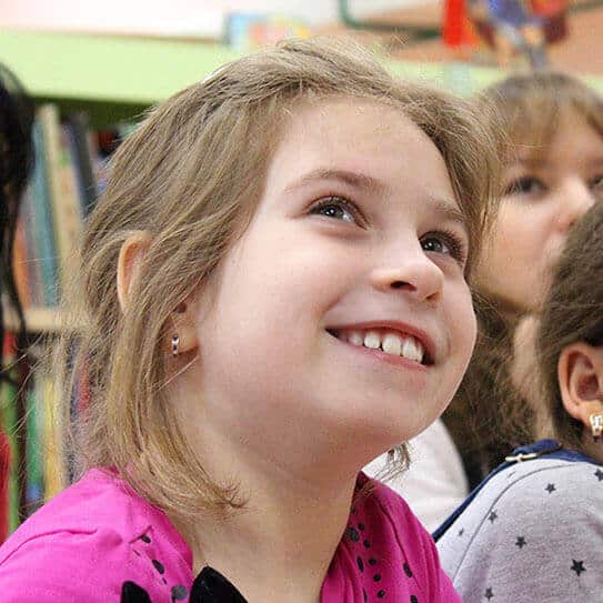 Young girl smiling brightly in classroom setting with other children visible in background.