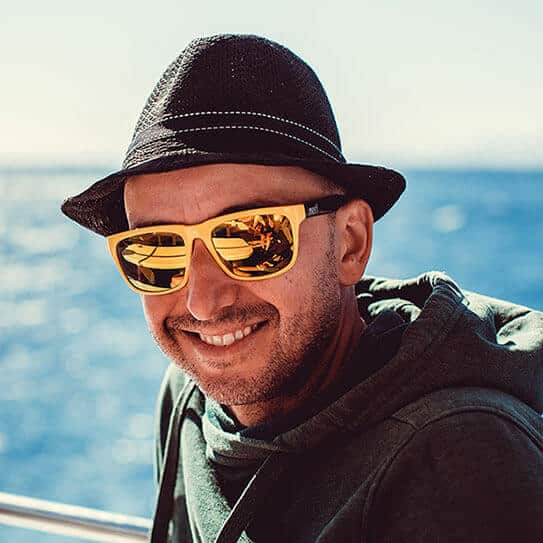 Man wearing hat and yellow sunglasses smiling on boat with ocean backdrop.