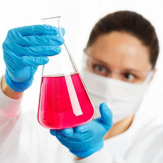 Lab technician in protective gear examining red liquid in beaker during dental research.