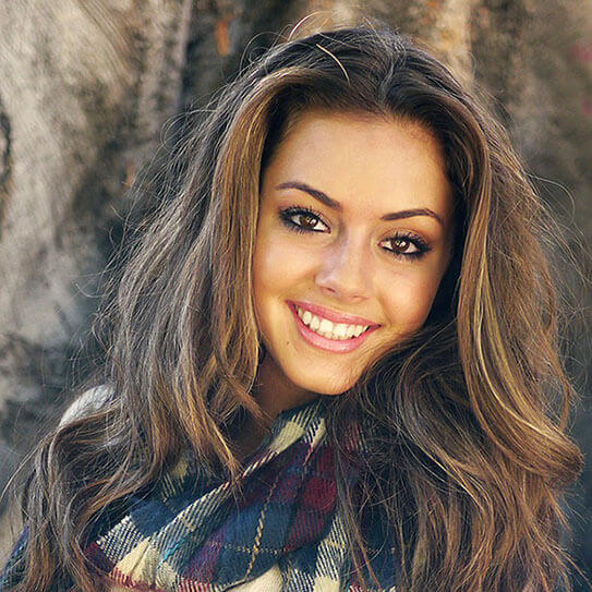 Young woman smiling brightly outdoors, wearing plaid scarf, professional headshot style portrait.