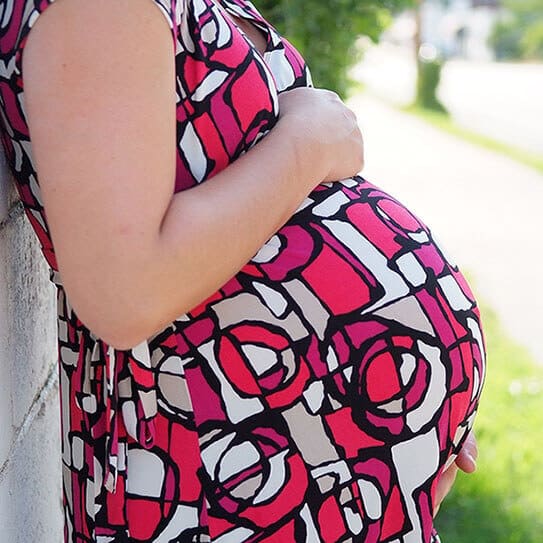 Pregnant woman in patterned dress cradling belly outdoors in natural daylight.