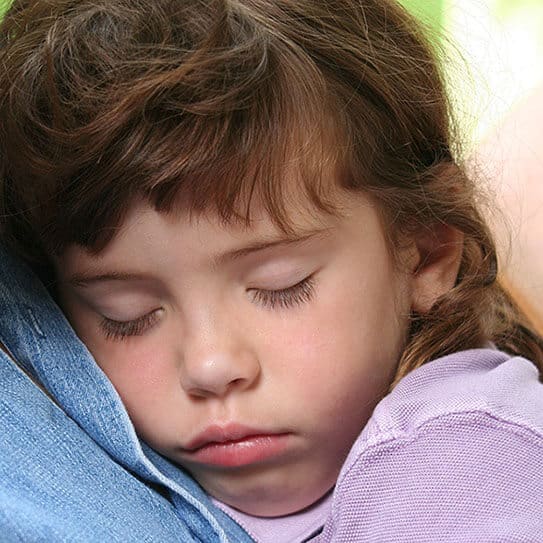 Young child sleeping peacefully, resting head on shoulder in casual indoor setting.