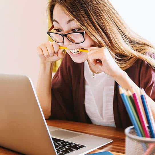 Young woman at desk with laptop holding colored pencils, appearing thoughtful or playful.
