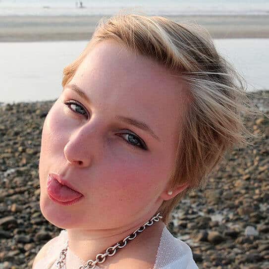 Woman with blonde hair smiling at camera on pebbly beach, wearing white shirt and chain necklace.