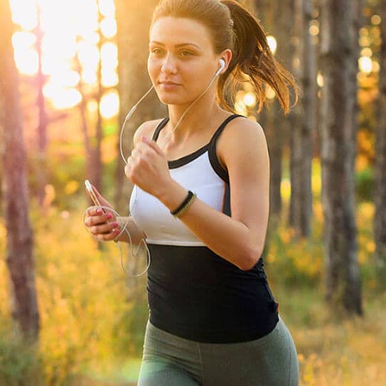 Woman jogging outdoors in forest at sunset, wearing athletic gear and smiling.