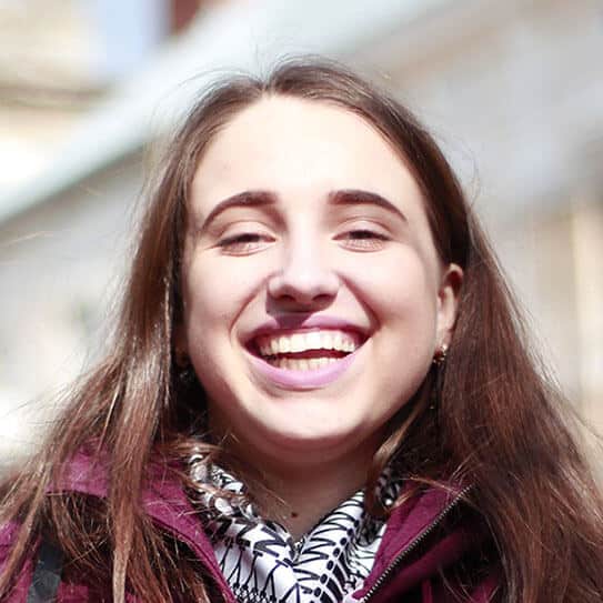 Smiling woman with long brown hair wearing purple jacket outdoors in daylight.