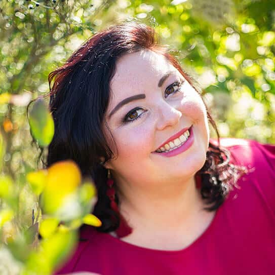Woman smiling outdoors among green foliage wearing magenta top