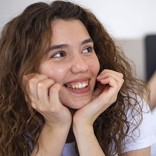 Happy woman with curly brown hair smiling, hands near face, in dental office setting.