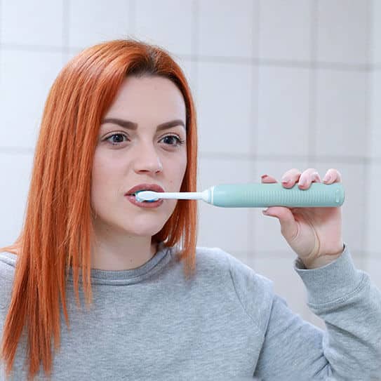 Woman with red hair brushing teeth with electric toothbrush in bathroom setting.