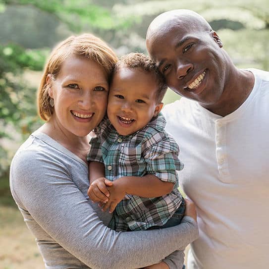 Happy family of three smiling outdoors in natural setting for dental practice website.
