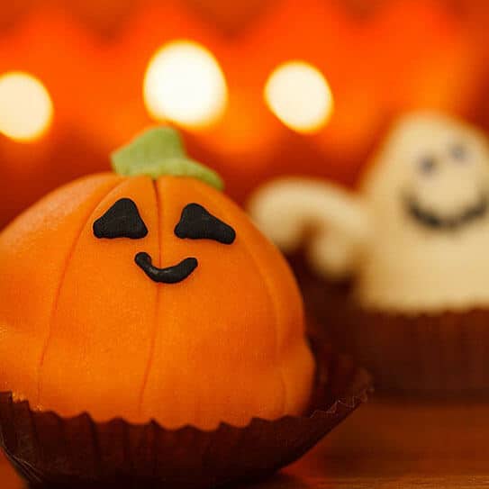 Smiling jack-o'-lantern pumpkin cake with Halloween decorations and glowing orange lights in background.