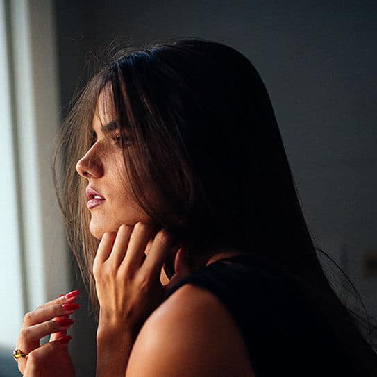 Woman with hand to face experiencing tooth pain near window with natural sunlight.