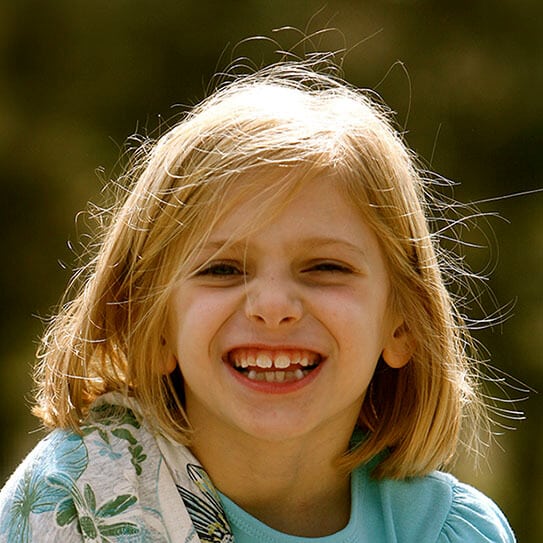 Young girl smiling brightly outdoors, displaying healthy teeth in natural sunlight.