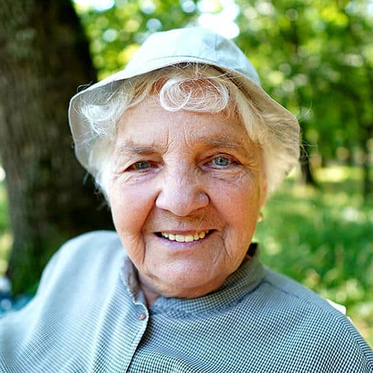 Smiling elderly woman wearing light blue shirt and white visor outdoors in park setting.
