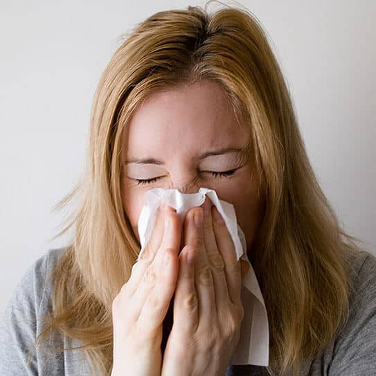 Woman with reddish-brown hair blowing nose into tissue against white background.