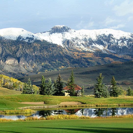 Scenic mountain landscape with snow-capped peaks, forested slopes, green meadows, and a rustic cabin by a pond.