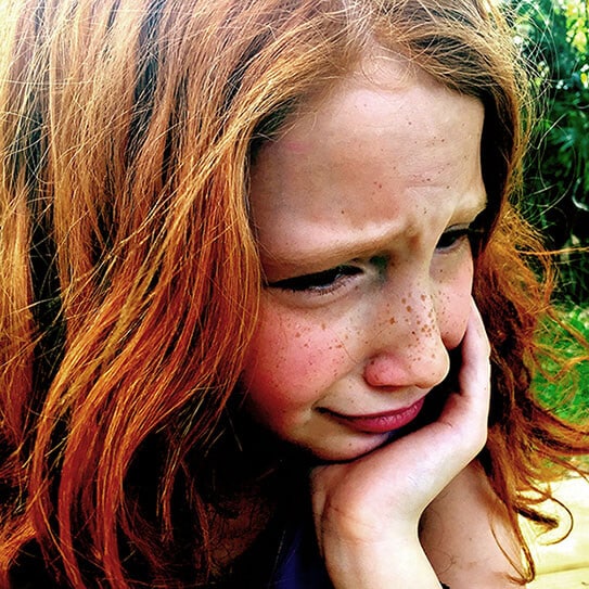 Young girl with reddish hair smiling thoughtfully outdoors, hand on chin.
