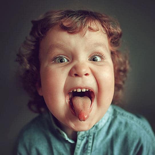 Young child with curly hair sticking out tongue playfully, wearing light blue shirt against neutral background.