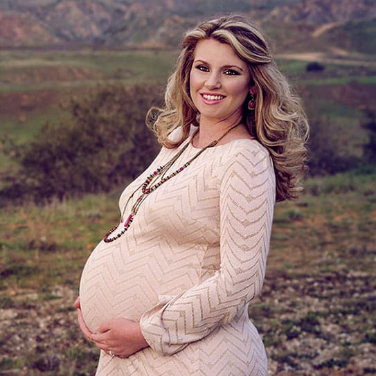 Pregnant woman in cream sweater posing outdoors with rural farmland background