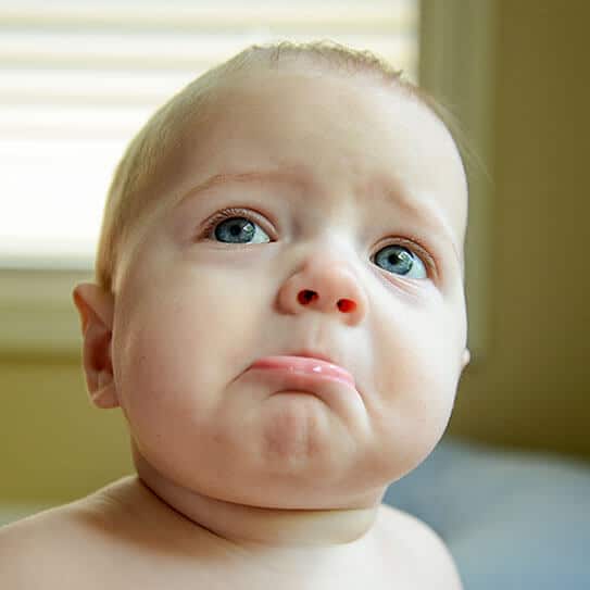 Baby with sad expression looking upward indoors, appearing upset or uncomfortable.