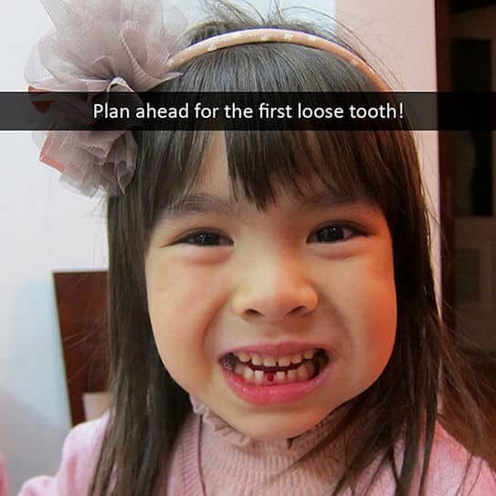 Young child smiling with loose tooth, wearing pink sweater and headband indoors.