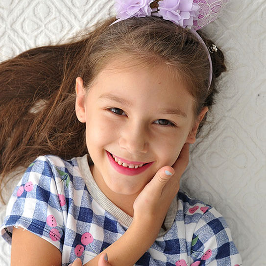 Young girl smiling brightly, wearing purple bow and plaid shirt, resting chin on hand indoors.