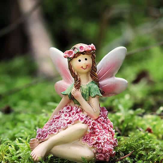 Fairy figurine with pink wings and flower dress sitting on moss in garden setting.