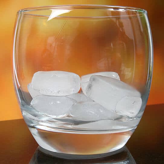 Glass of water with ice cubes on wooden surface, warm lighting background.