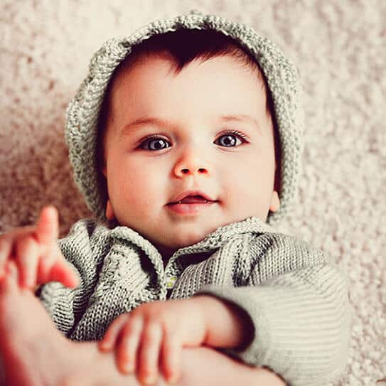 Smiling infant wearing gray knit hat and sweater against neutral beige background.