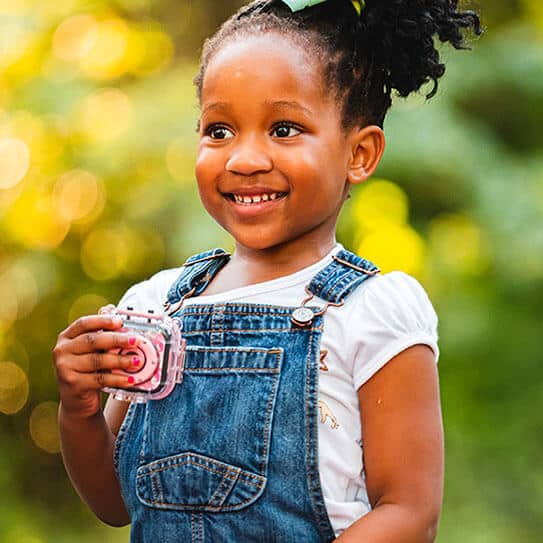 Young child in denim overalls smiling outdoors while holding pink cupcake.