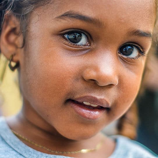 Young child with bright eyes smiling gently at camera in outdoor setting.