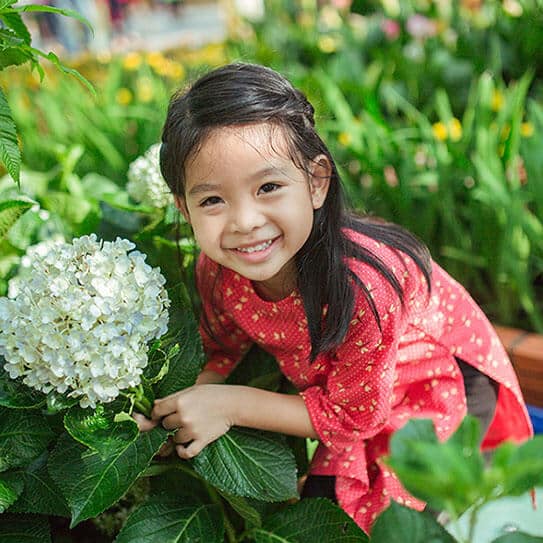 Young girl smiling at camera while holding white flower in garden setting.