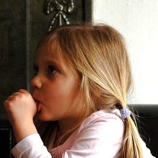 Young girl with blonde hair in pink shirt, resting chin on hand, contemplative expression indoors.