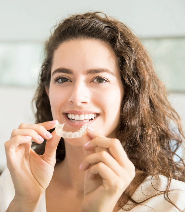 A woman with curly hair holding a clear aligner and smiling at Denver Orthodontics and Dental Lounge in Denver CO