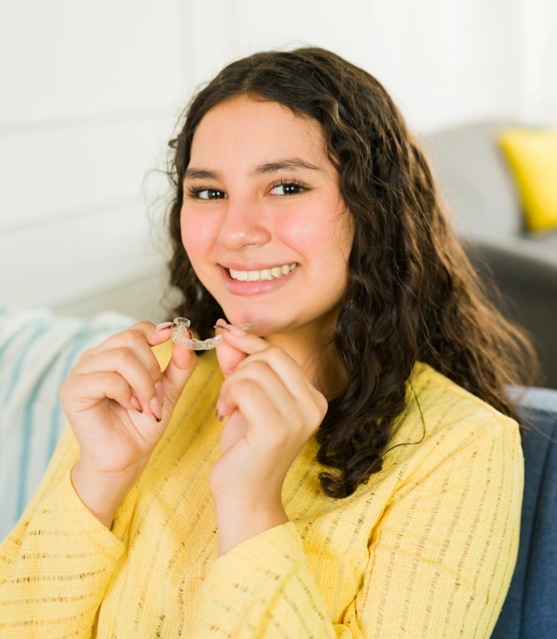 A teenage girl in yellow holding a clear aligner and smiling at Denver Orthodontics and Dental Lounge in Denver CO