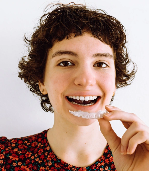 A young woman with curly hair holding a clear aligner and smiling at Denver Orthodontics and Dental Lounge in Denver CO